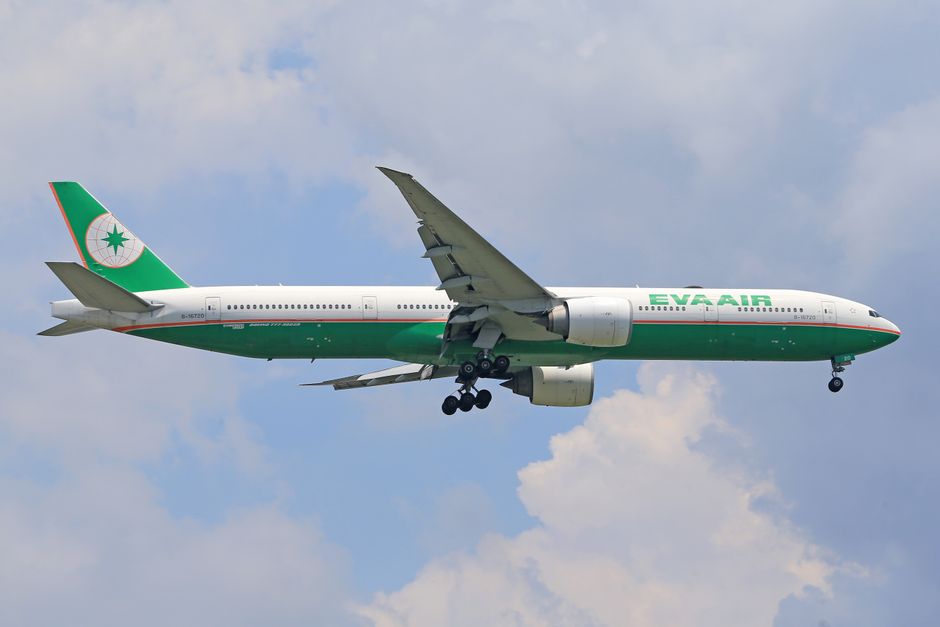 A large EVA Air commercial aircraft is captured mid-flight against a partly cloudy sky, displaying a white fuselage with green and red accents. The aircraft's wings extend outward, with visible flaps 