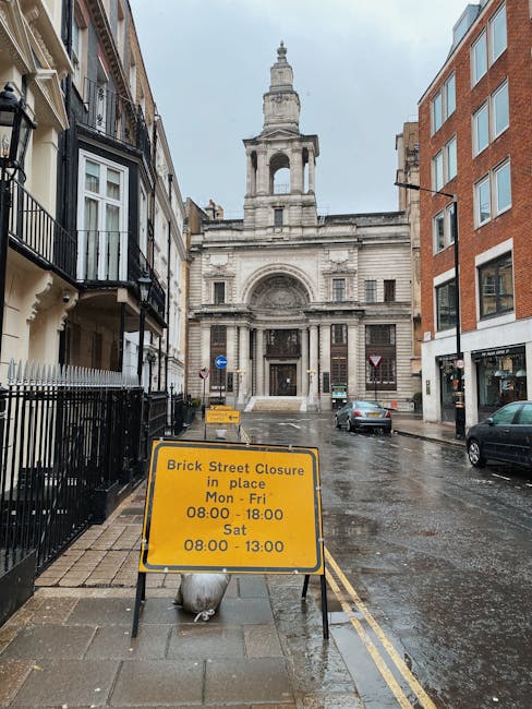 Photograph of a narrow urban street in Hammersmith, W6, showing a yellow road closure sign placed on the wet sidewalk in front of a historic stone building with a central arch and a tower-like structu