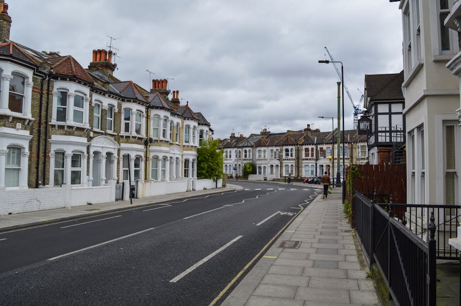 Photograph of a narrow urban street in Hammersmith, W6, showing a yellow road closure sign placed on the wet sidewalk in front of a historic stone building with a central arch and a tower-like structu